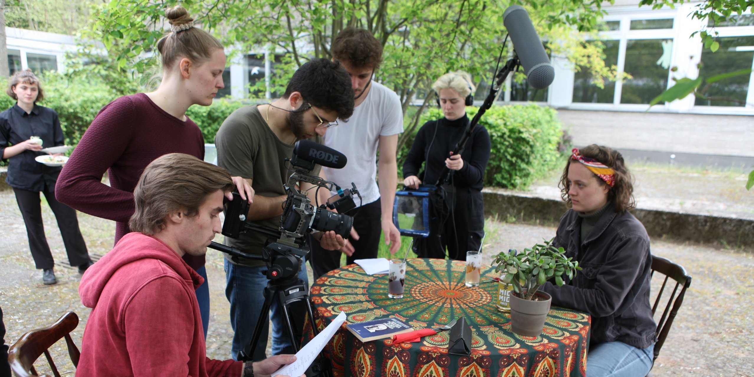 zwei Personen sitzen sich an einem Tisch im Freien gegenüber, neben dem Tisch steht ein Filmteam