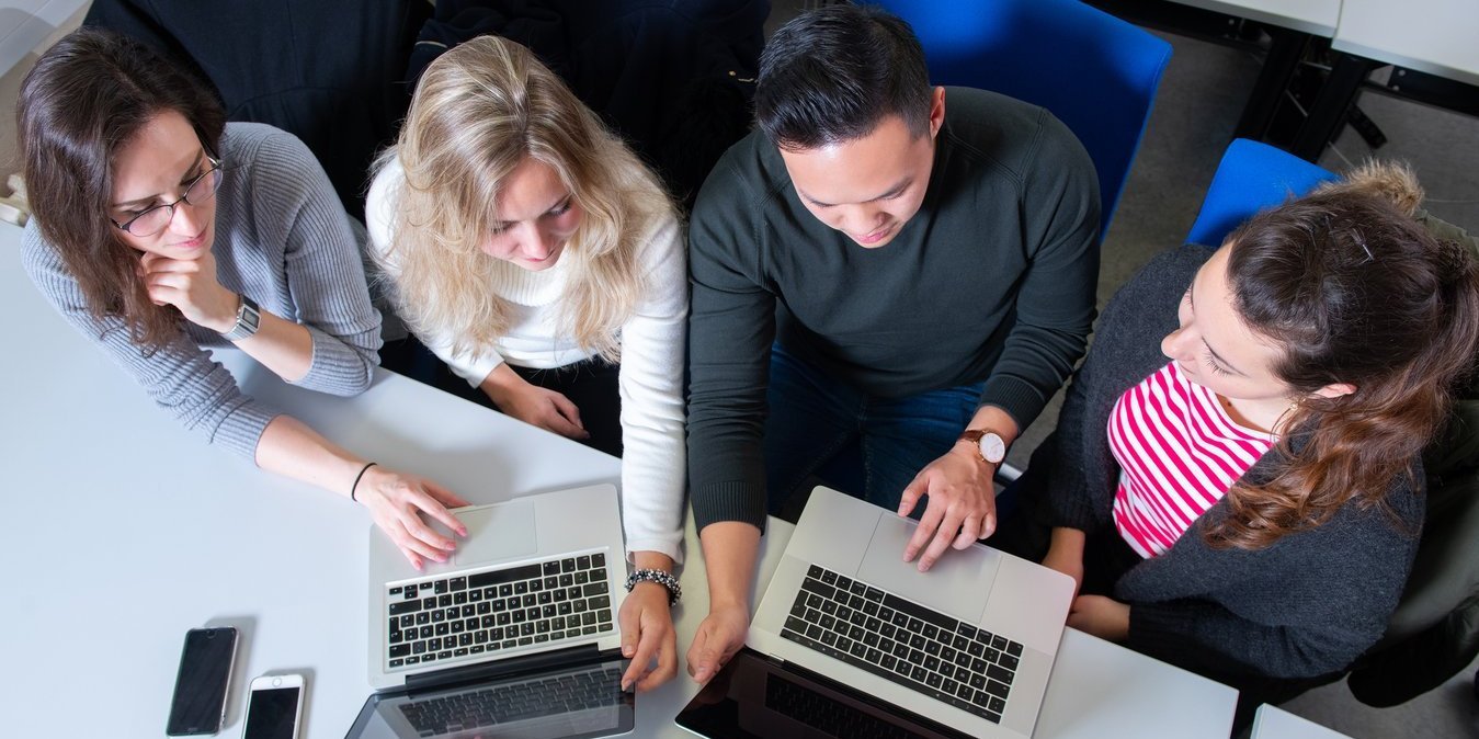 Vier Studierenden sitzen nebeneinander an Tischen und schauen auf zwei Laptops.