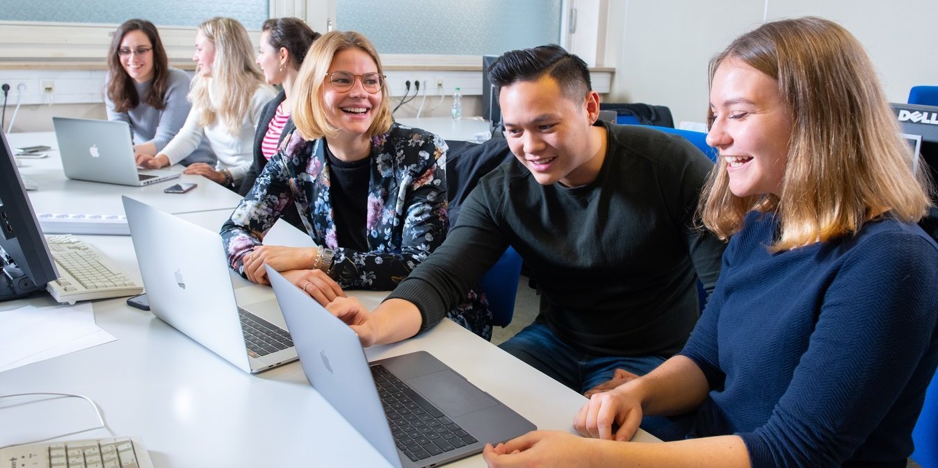 Mehrere Studierende sitzen nebeneinander an Tischen vor ihren Laptops und besprechen etwas.